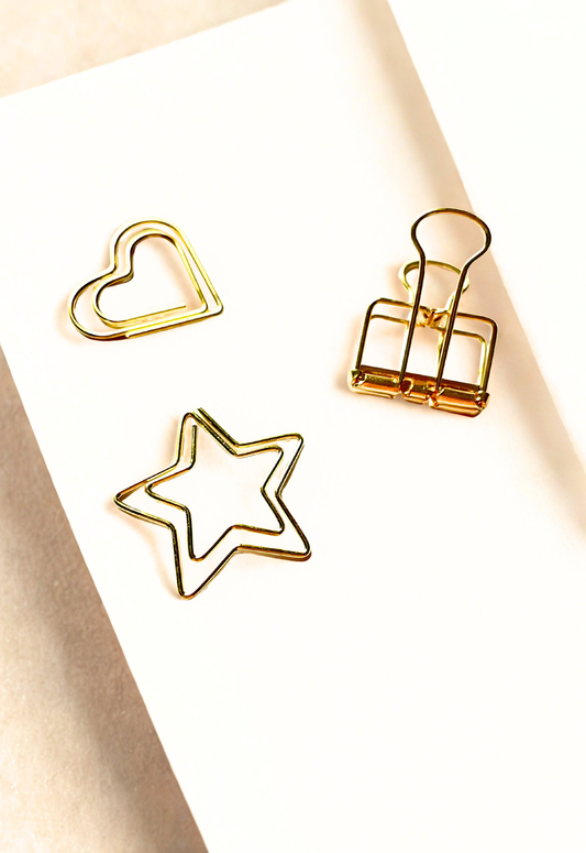 A flat-lay of stationery items on a light gray background: a lavender pen, a sheet of rectangular sticky notes in pastel shades from light purple to blue, and gold metal paperclips in binder, heart, and star shapes.