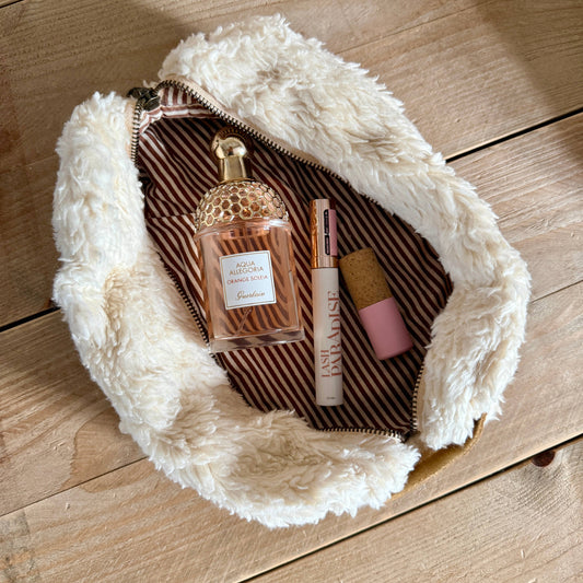 A fluffy cream faux-fur zip pouch with a tan leather handle embroidered with the word 'Liebe' in black script, shown open on a light wood surface. Inside are several cosmetic items, including a perfume bottle and lip product.