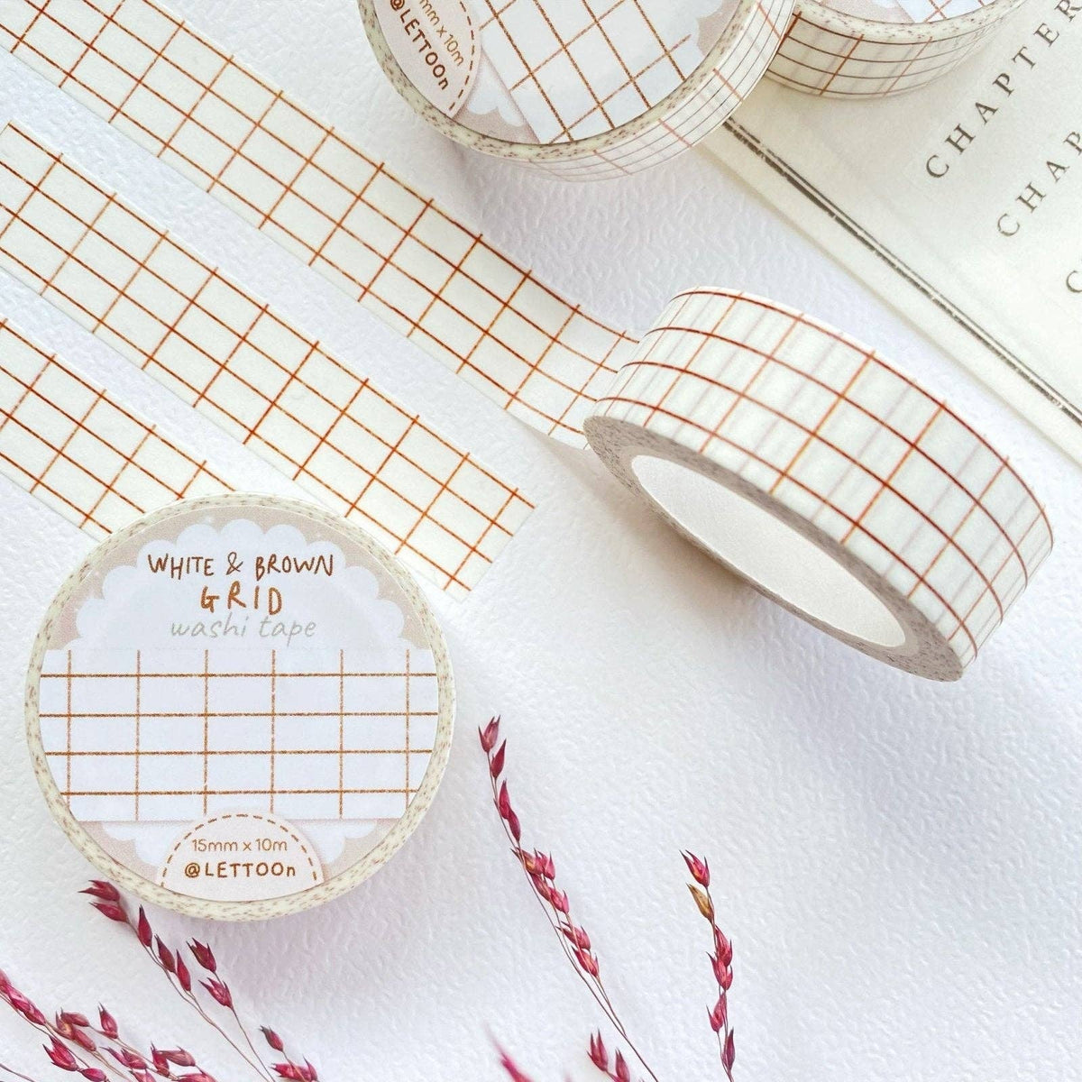 White washi tape with a brown grid pattern, shown in a roll with a paper label and unrolled on a white textured surface next to a book and dried grass stems.