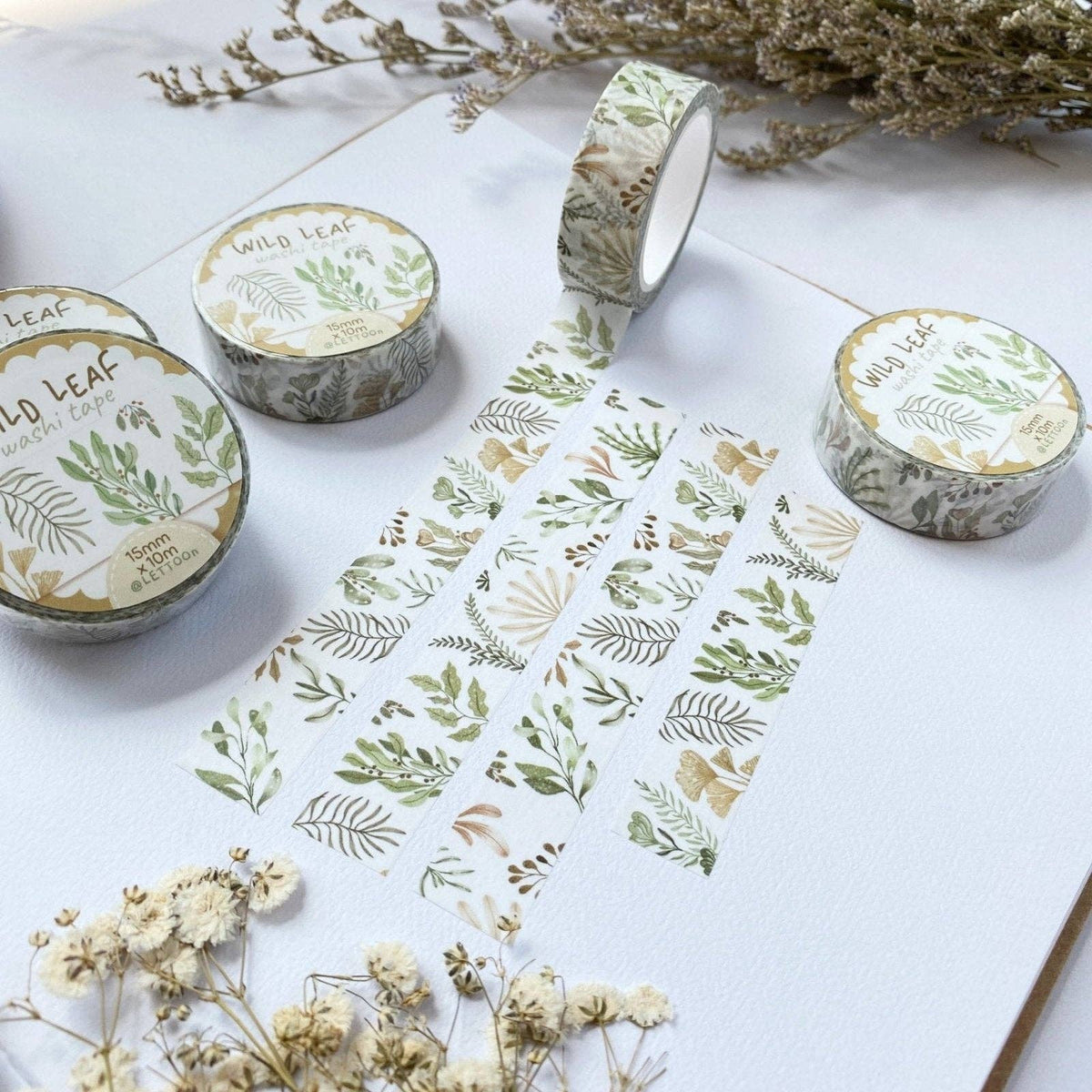 Three rolls of washi tape with a botanical leaf pattern are arranged on a white textured surface with dried flowers. The tape features green, beige, and brown leaves on a white background.