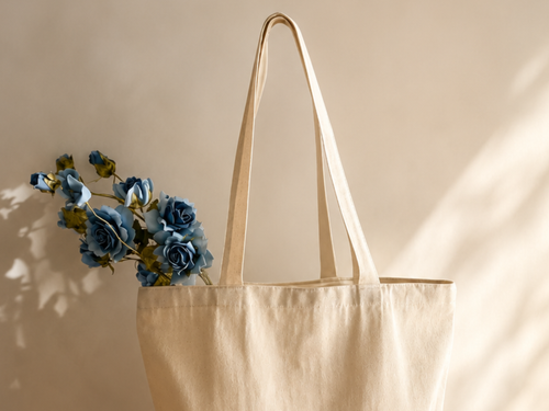 A natural beige canvas tote bag hanging against a light wall, with blue artificial roses tucked into one of its handles.
