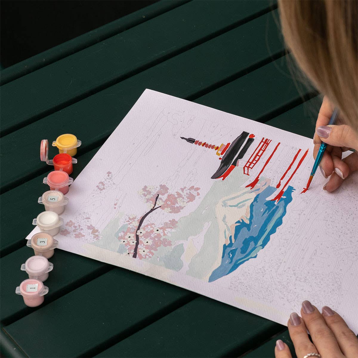 Person painting a scenic artwork with watercolors on a green table.