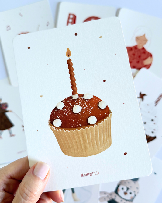 A square art print or card featuring a decorated cupcake with a single lit candle on a white background, with small scattered star-like specks and the text 'PAPIEROSTIQUE.FR' at the bottom.