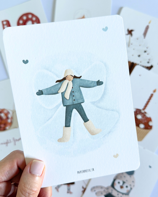 A vertical art print featuring a watercolor illustration of a child lying down in a snow angel pose, wearing a blue-gray coat, matching pants, and beige boots. The print has a white border and is displayed against a clean white background.