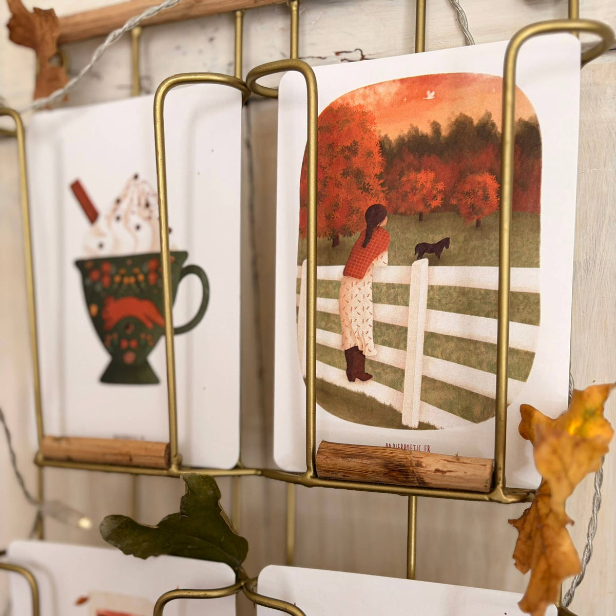 A set of illustrated greeting cards displayed in a gold metal wire rack. The featured card on the right shows an autumn scene with a woman by a white fence and a dog, while the card on the left shows a patterned mug with a hot drink.