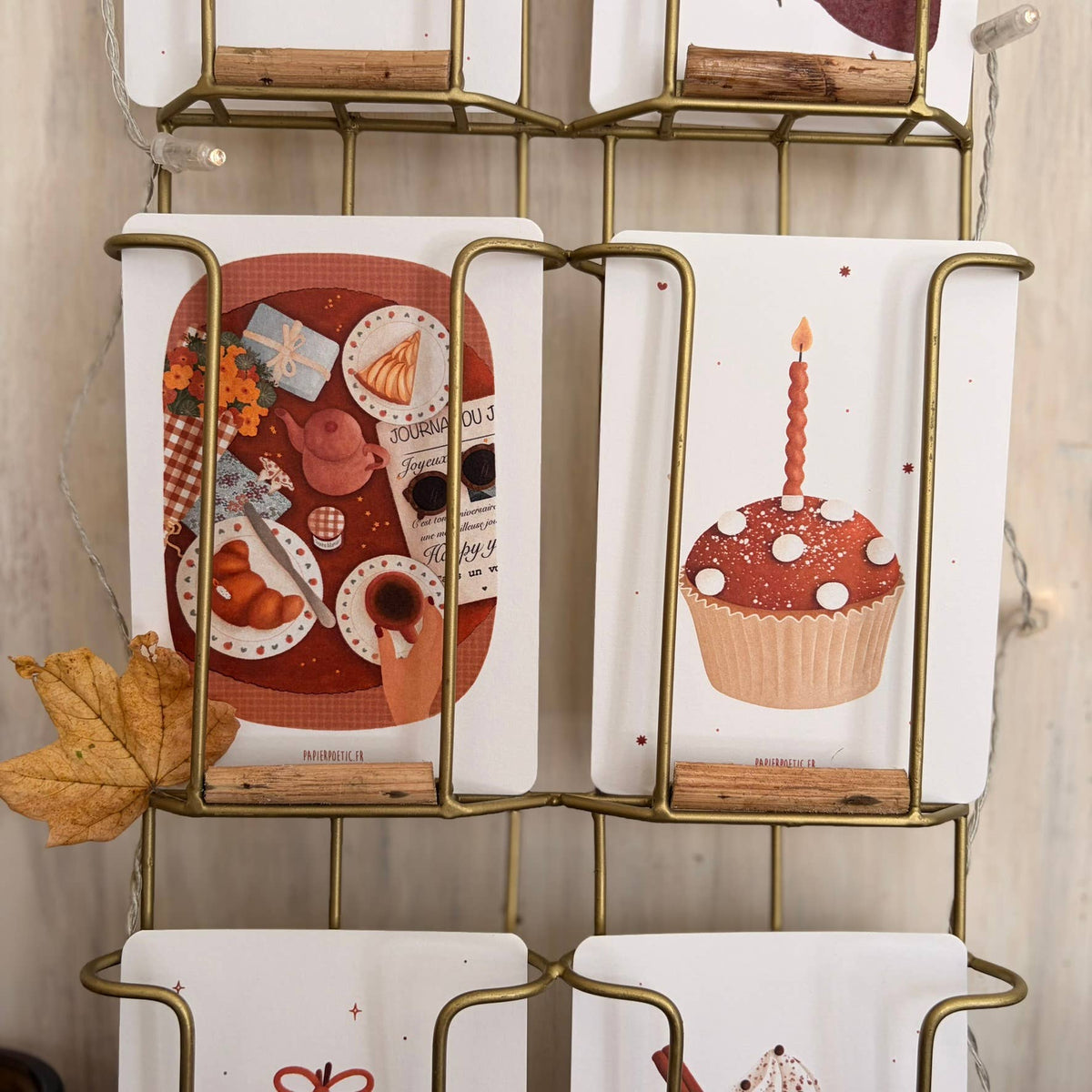 A brass wire hanging rack with white fabric cloths featuring printed autumn-themed illustrations, including a breakfast scene, a cupcake, and a ribbon bow, displayed against a light wood wall.