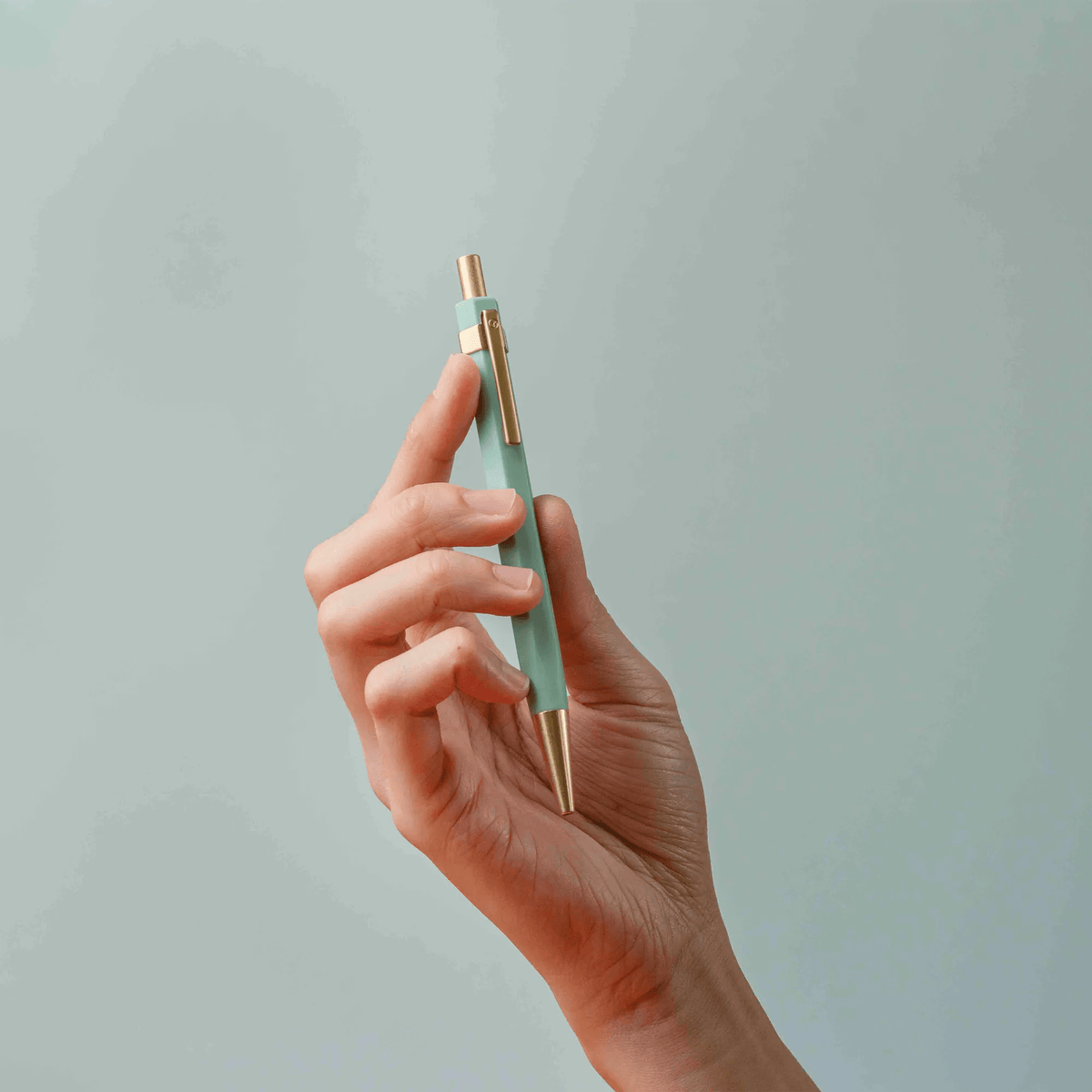 Hand holding a mint green pen with gold accents against a light gray background
