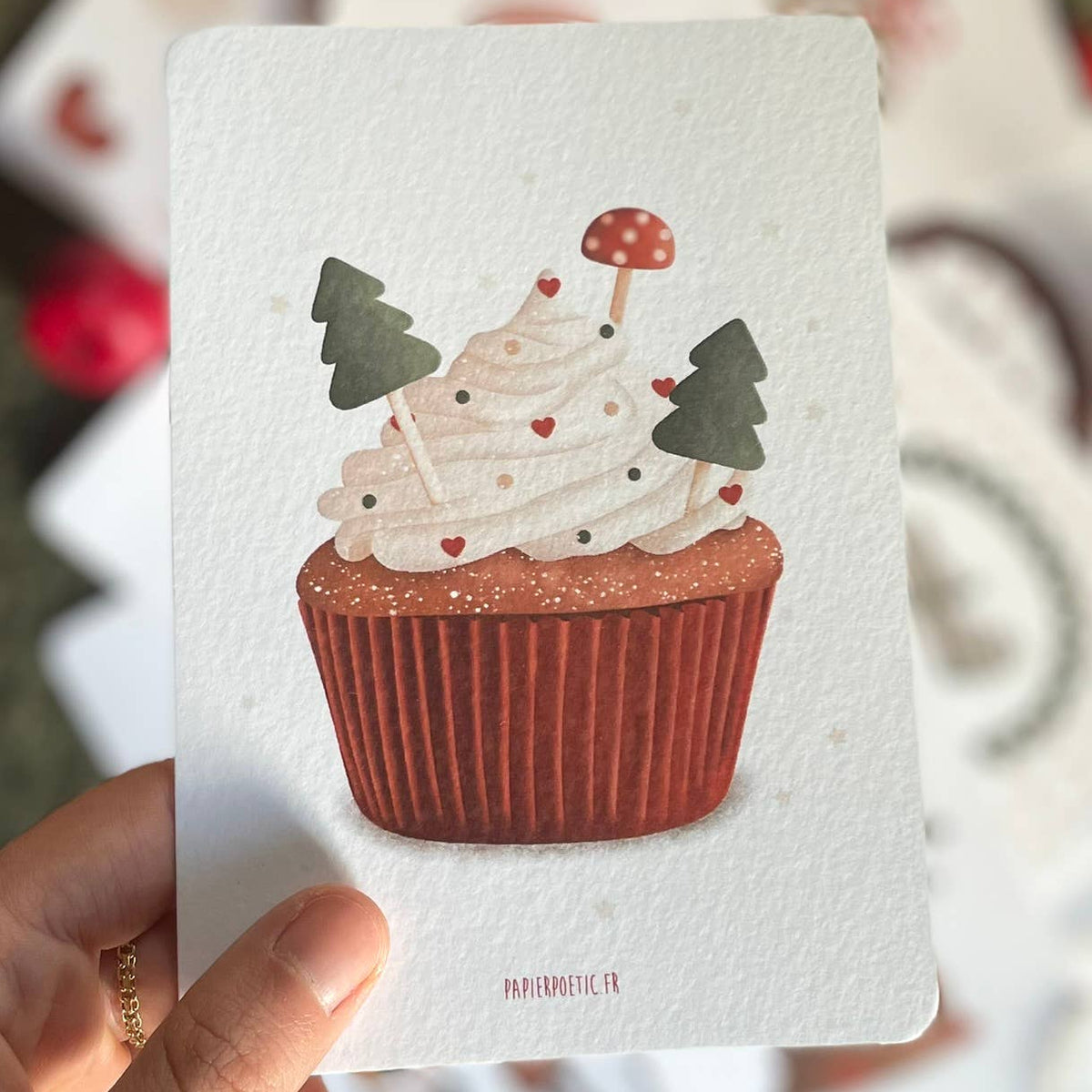 A hand holds a square art print featuring a watercolor-style cupcake with white frosting, red berries, green trees, and a small red-and-white mushroom topper. The print has the visible brand text 'MAMAMOOE.
