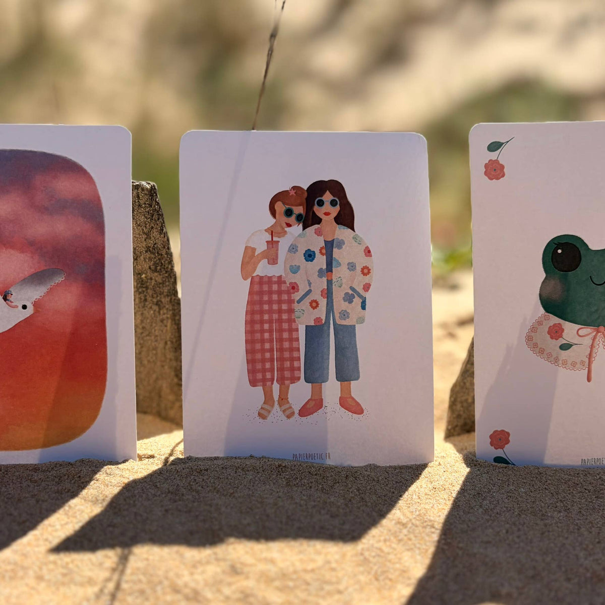 Three illustrated art cards are propped upright in sandy ground outdoors. The center card shows two women in colorful outfits, with blurred natural scenery behind them.