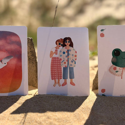 Three illustrated art cards are propped upright in sandy ground outdoors. The center card shows two women in colorful outfits, with blurred natural scenery behind them.