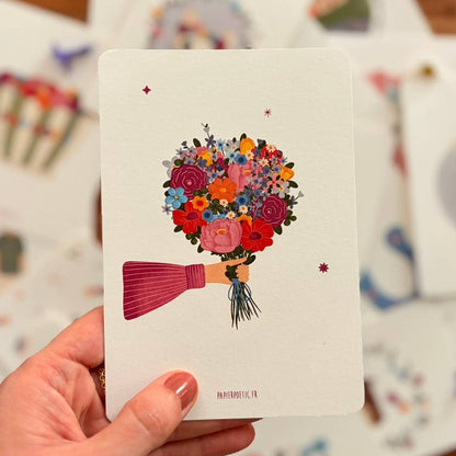 Hand holding a card with a colorful bouquet illustration on a wooden surface with more cards.