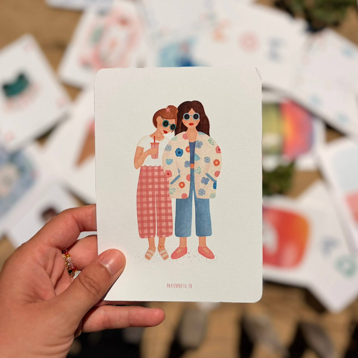 A hand holds a white art print card featuring an illustration of two women in colorful outfits. The background is a soft-focus display of other art prints.