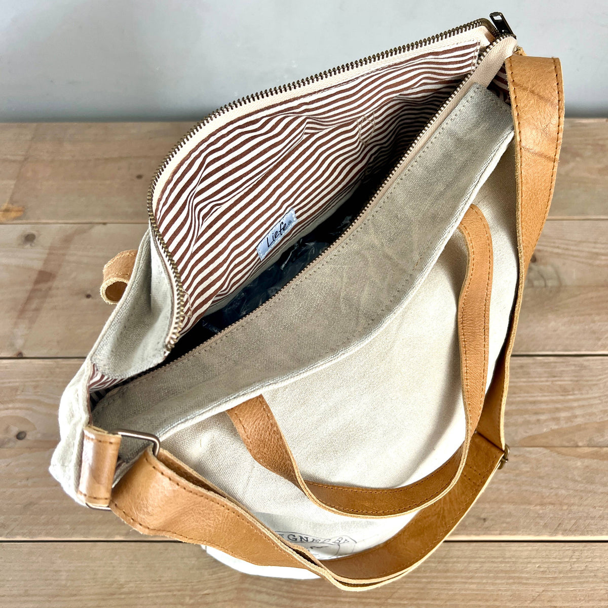 An open canvas shoulder bag with tan leather straps and a striped fabric lining, photographed from above on a wooden surface.