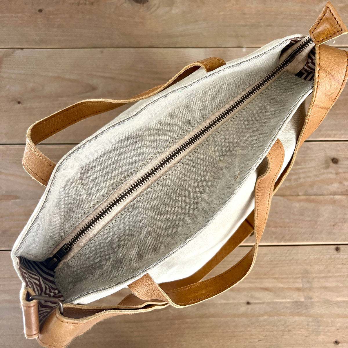 An open canvas tote bag with tan leather handles and trim, showing a light gray interior lining and a zipper, placed on a rustic wooden plank surface.