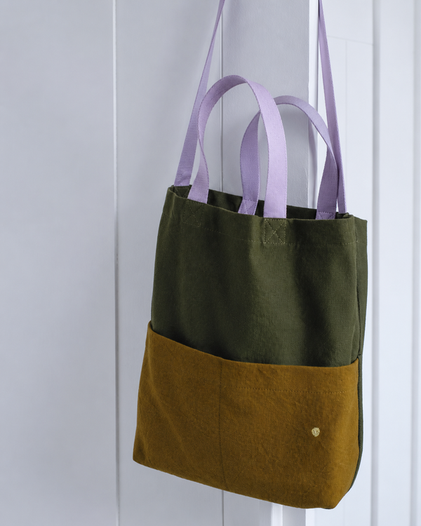 A two-tone fabric tote bag hanging against a white paneled door. The upper half is olive green, the lower half is mustard yellow, and the shoulder straps are light purple.