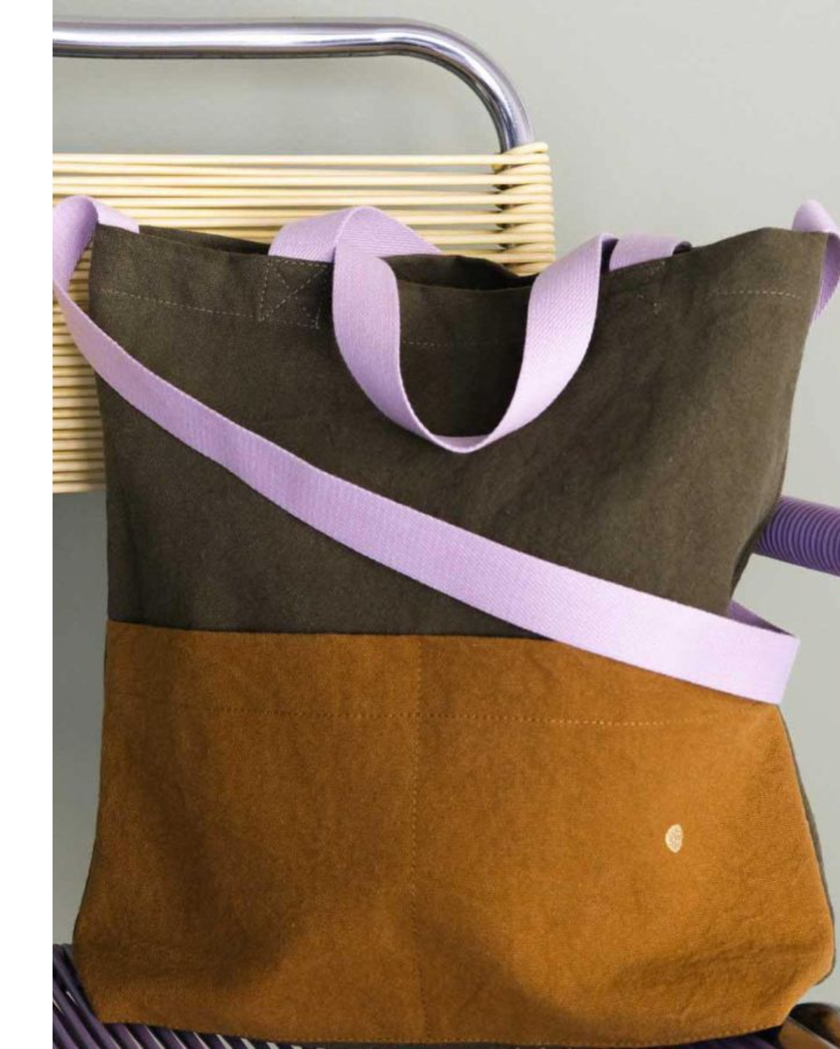 A large tote bag made of two-tone fabric, featuring a dark olive green top half, a mustard brown bottom half, and light purple handles and strap, resting on a wicker chair.