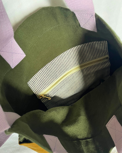 Close-up of the interior of an olive green tote bag, showing a zippered pocket with a striped lining and lavender fabric handles.