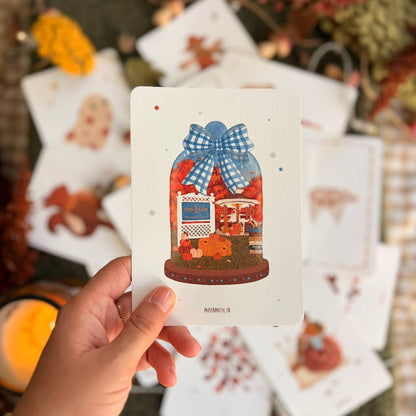 A hand holds a white art print card featuring an illustration of a glass jar filled with autumn leaves and a blue gingham bow, with other blurred art prints scattered in the background.