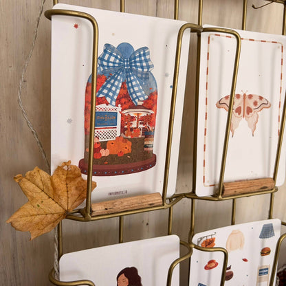 A gold metal wall-mounted card holder displaying illustrated cards. The cards feature whimsical designs including a blue gingham bow, a carousel, a butterfly, and small food illustrations.
