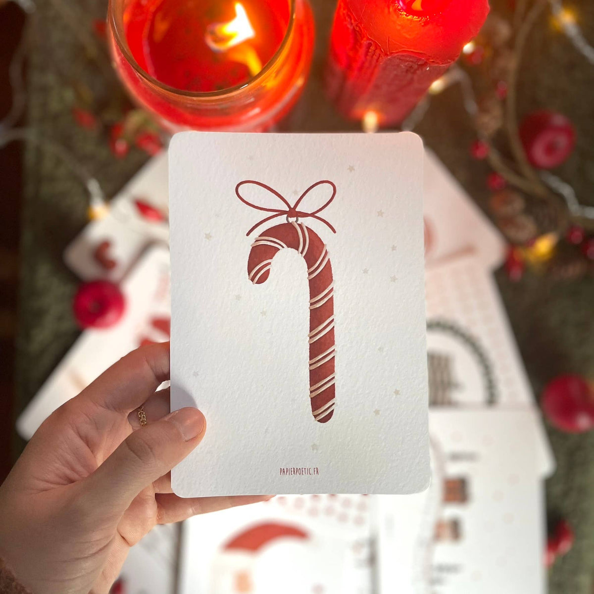 A hand holds a white textured Christmas card featuring a red candy cane with a bow, with blurred red candles and holiday decorations in the background.