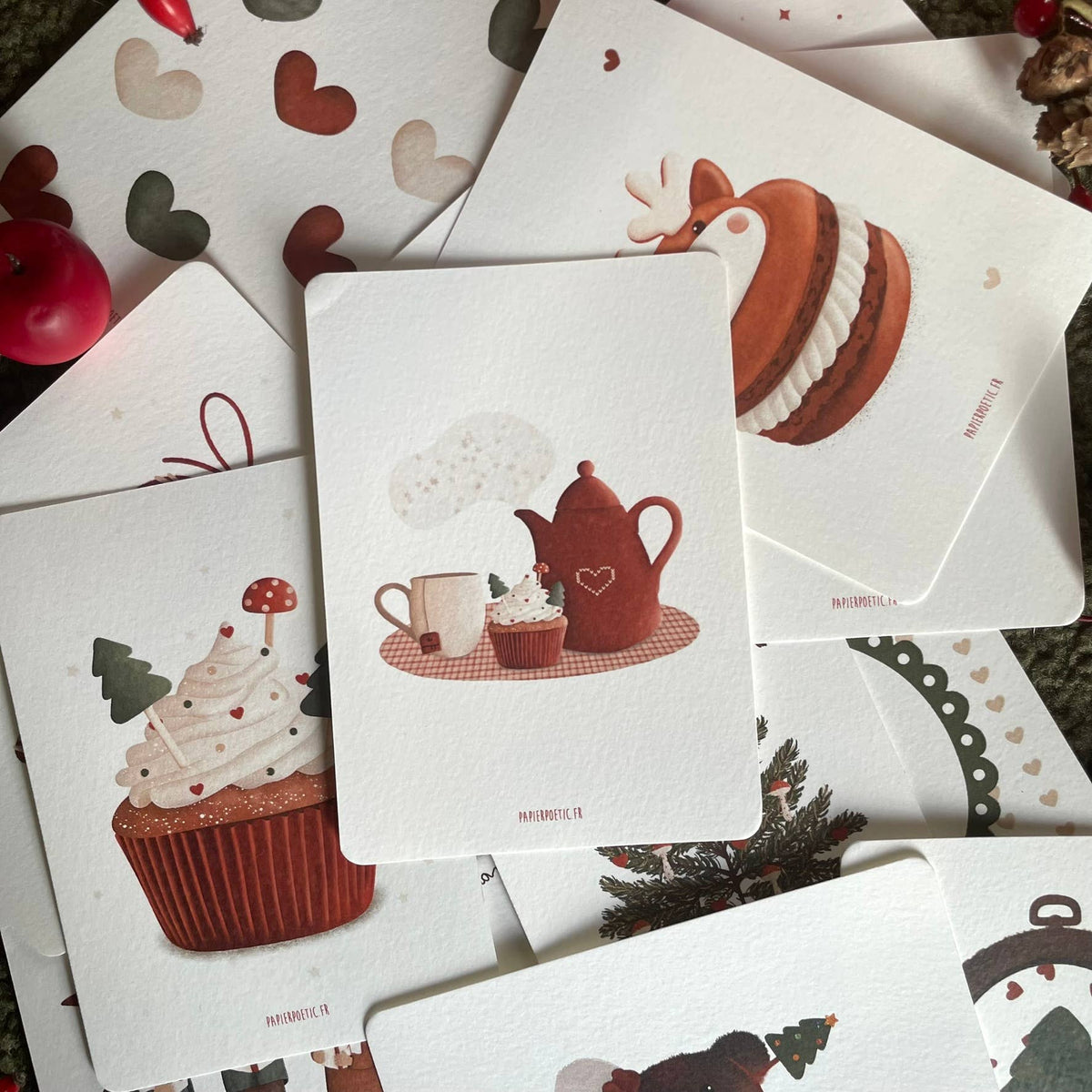 A flat-lay arrangement of white greeting cards featuring winter and holiday illustrations, including a central card with a red teapot, cup, and cupcake, surrounded by other cards with cupcakes, a reindeer cake, and heart motifs.