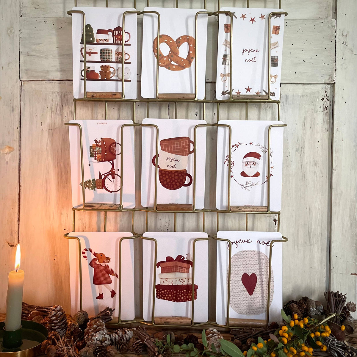 A set of nine white kitchen towels with various holiday and coffee-themed illustrations, displayed on a gold metal rack against a rustic white wood wall with a lit candle and pinecones in the foreground.