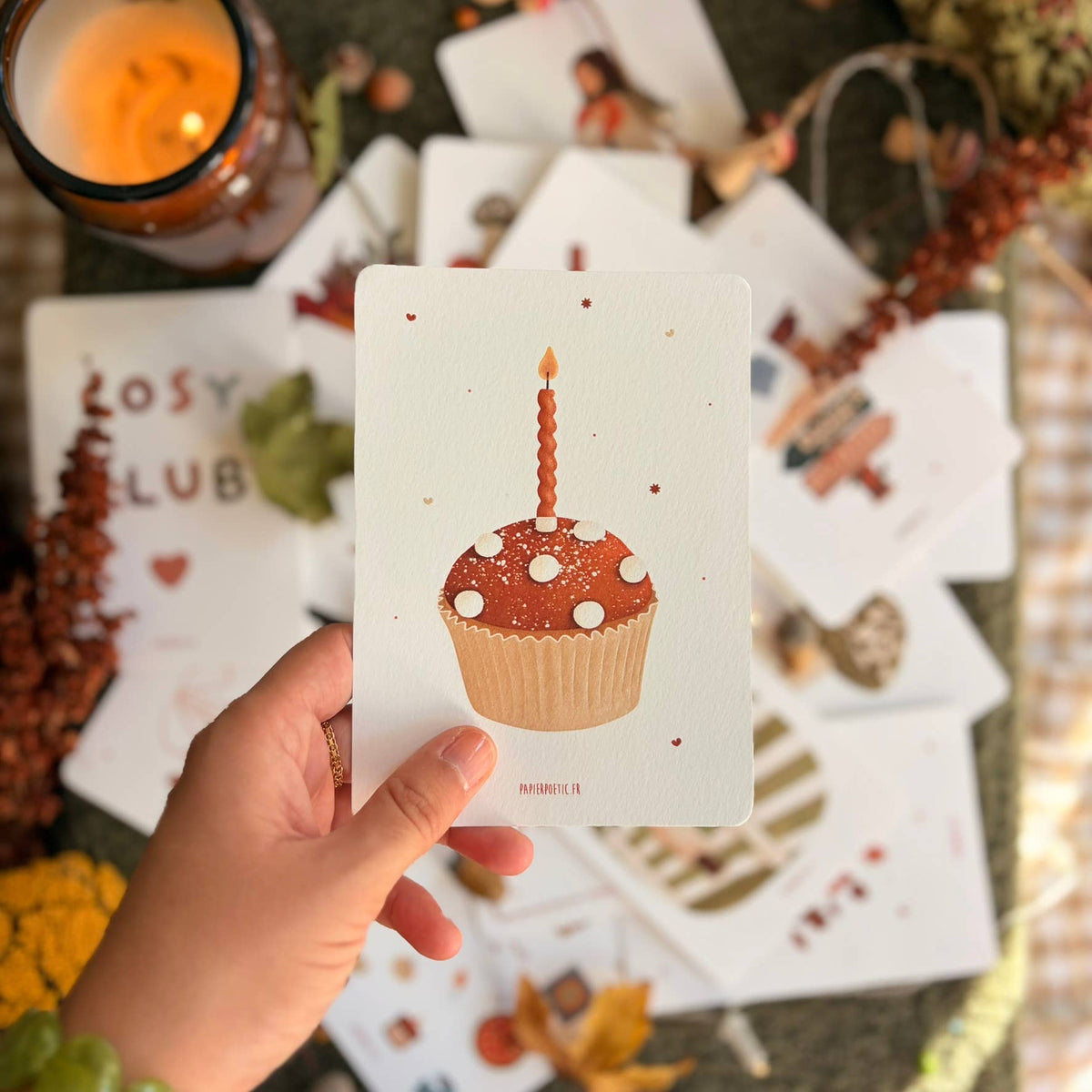 A hand holds a white art print card featuring a cupcake illustration, surrounded by scattered art prints, a lit candle, and autumn-themed decorative elements.
