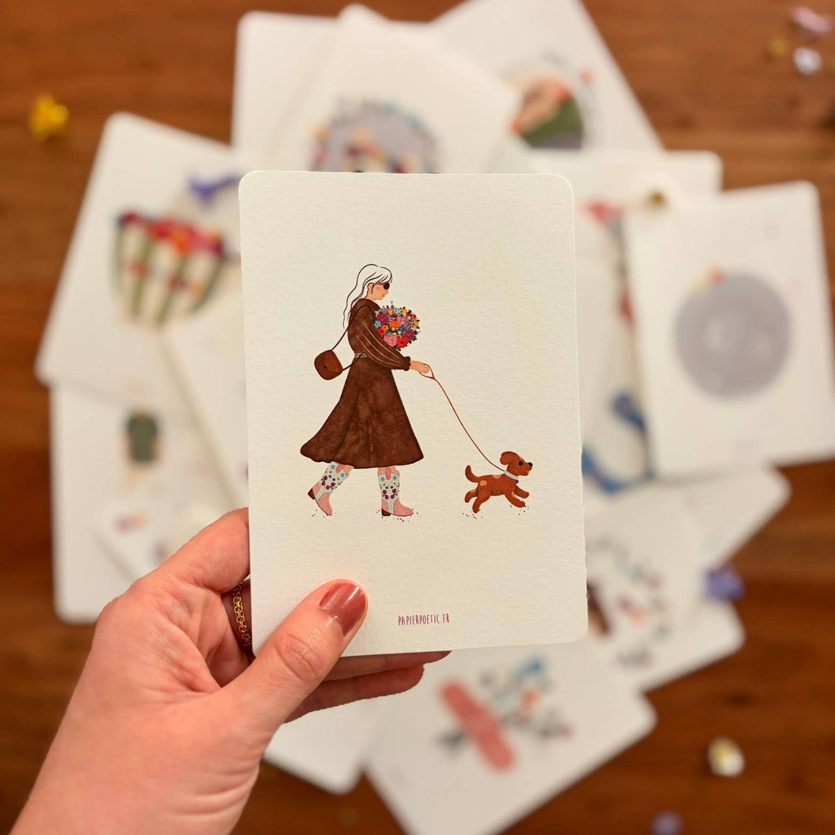 A hand holds a white art print card featuring an illustration of a woman in a brown coat walking a small brown dog while holding a colorful bouquet. A scattered pile of similar art cards lies on a wooden surface in the background.