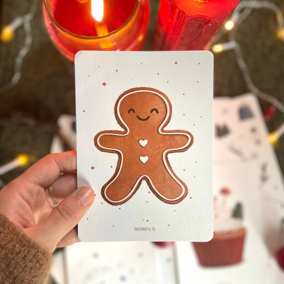 A hand holds a white greeting card featuring a brown gingerbread man with small heart details, set against a cozy holiday background with lit red candles and warm fairy lights.
