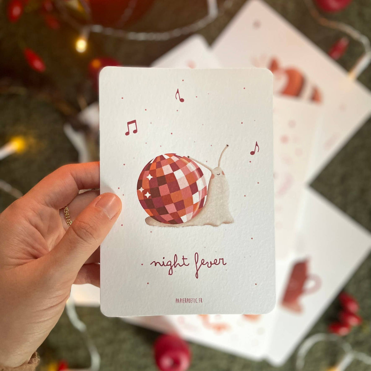 Hand holding a card with a checkered snail illustration and 'night fever' text, with blurred Christmas lights in the background.