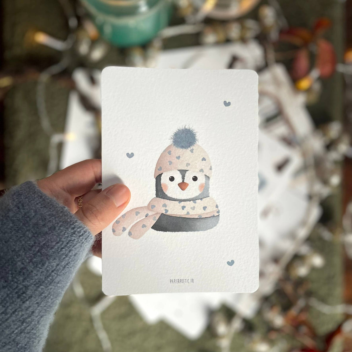 A hand wearing a gray sweater holds a white art print featuring a cute penguin wearing a beige and gray patterned winter hat and scarf. The background is a cozy, out-of-focus holiday scene with warm lights and decorations.
