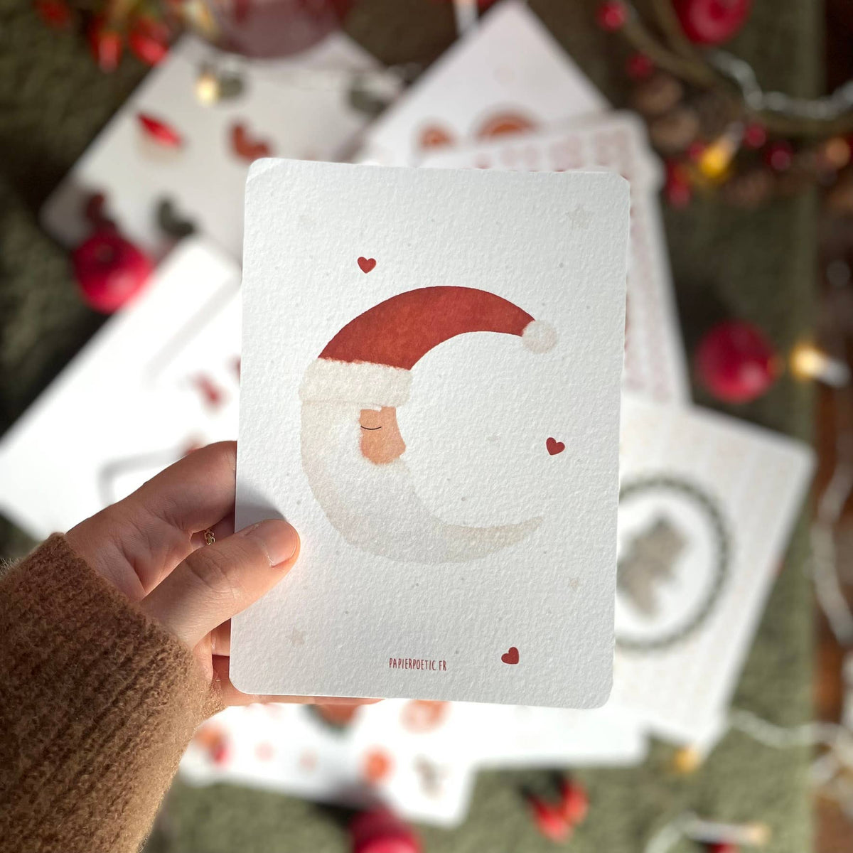 A hand wearing a brown knit sleeve holds a square white card featuring a watercolor-style Santa Claus face inside a crescent moon with small red hearts. Other similar cards are scattered in the background on a dark surface with blurred holiday lights and red ornaments.