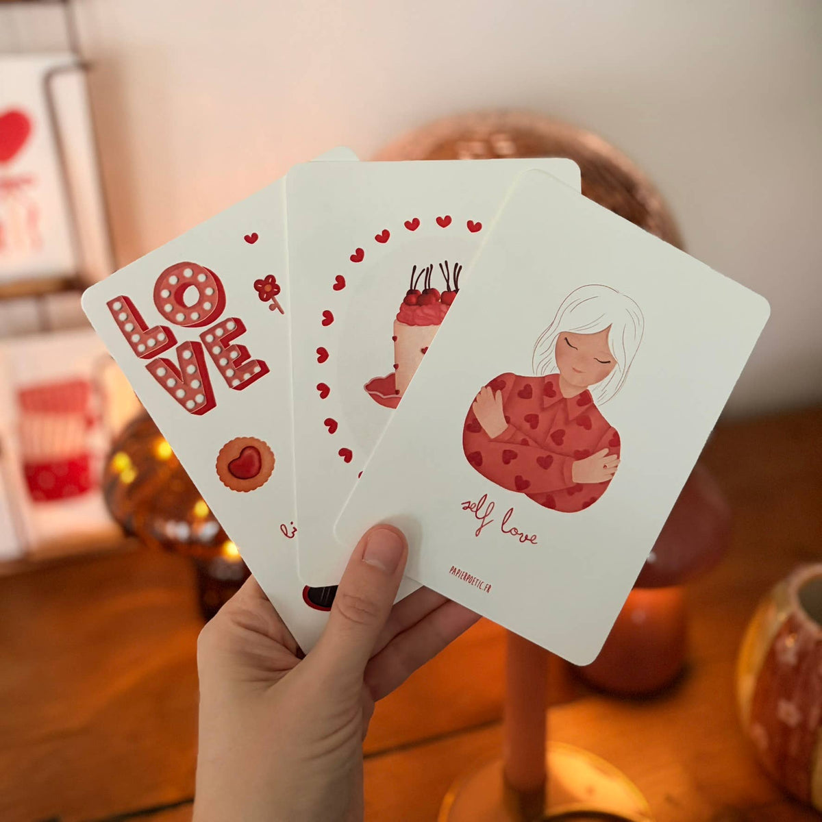 A hand holds three illustrated cards fanned out in a warm indoor setting. The cards feature red and pink Valentine-themed artwork, including the word "LOVE," a cupcake with hearts, and a woman with the words "all love.