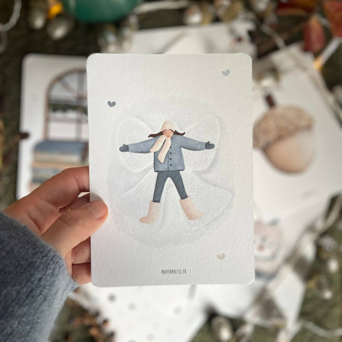 A hand holds a white art print card featuring a whimsical illustration of a child dressed in winter clothes, standing in the shape of a snow angel. The background is a soft-focus outdoor scene with natural elements.