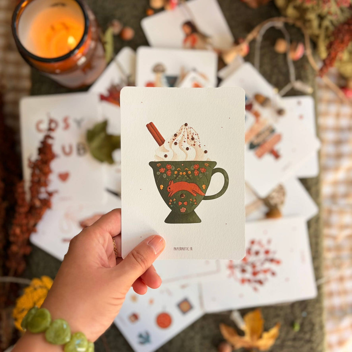 A hand holds a white art print card featuring a whimsical illustration of a green mug filled with whipped cream, cocoa powder, and a cinnamon stick. The background is a styled flat-lay of other art prints, a lit candle, and dried autumn foliage.