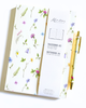 A Kantos Taccuino A5 notebook with a cream cover decorated with colorful wildflowers, shown with its white branded dust jacket and a matching floral gold pen on a white background.