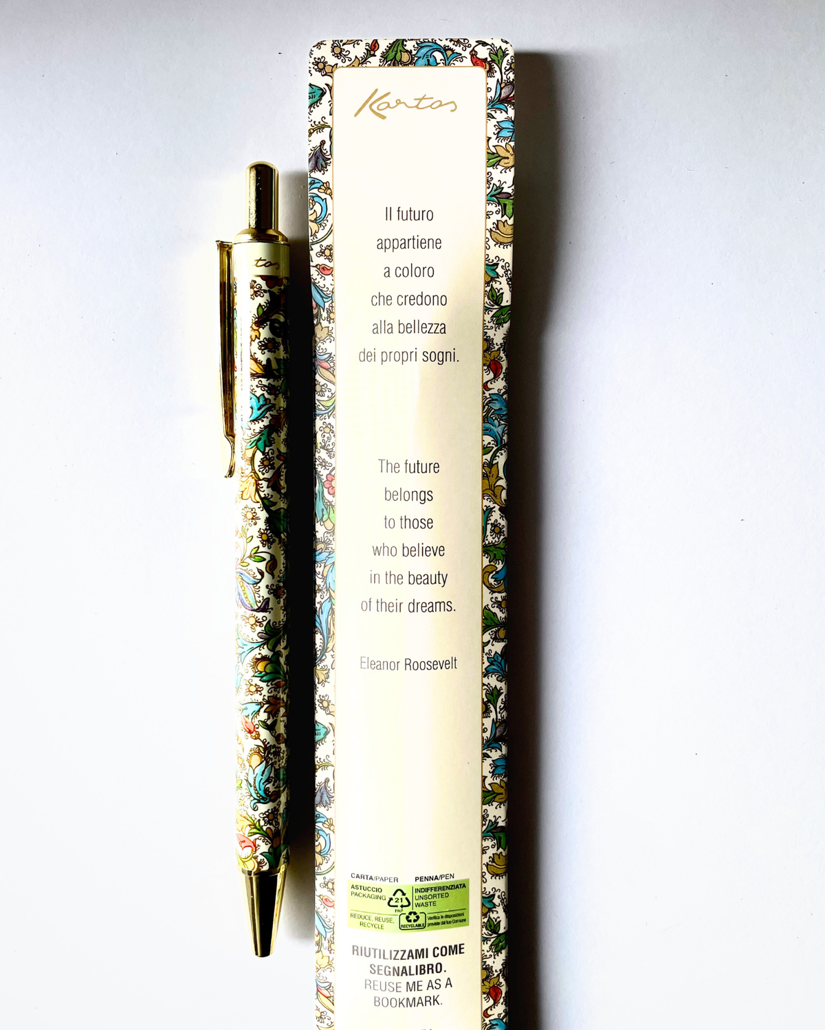 A gold-plated ballpoint pen with ornate multicolor floral enameled detailing, displayed next to its cream-colored packaging box featuring Italian and English text and a quote by Eleanor Roosevelt.