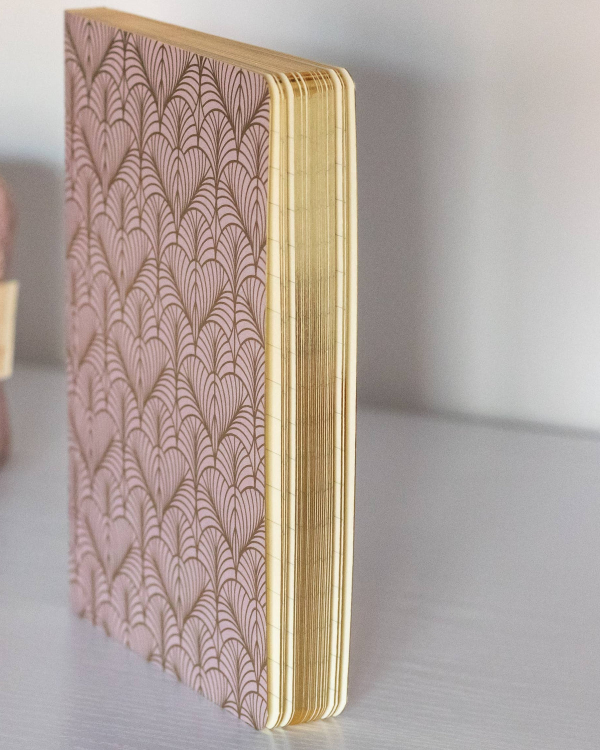 A pink patterned notebook with gold-toned edges stands upright on a light gray surface, next to a matching pink patterned pouch and a gold-and-white pen.