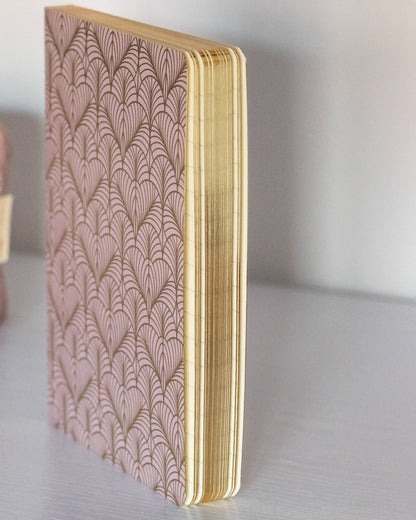 A pink patterned notebook with gold-toned edges stands upright on a light gray surface, next to a matching pink patterned pouch and a gold-and-white pen.