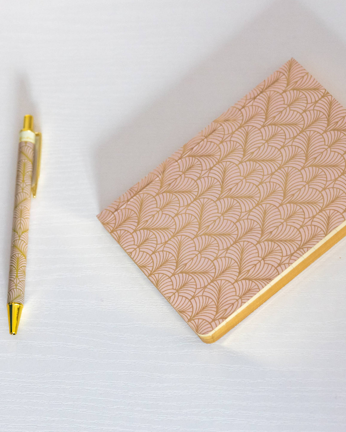 A peach and gold patterned notebook with a gold pen and a matching pink and gold patterned pouch on a white surface.