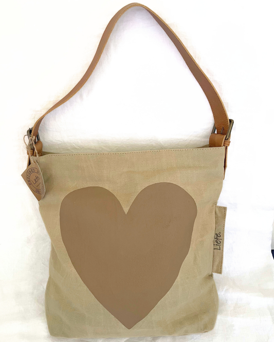 A canvas shoulder bag in warm off-white with a large muted tan heart printed on the front, hanging against a brown garment. A small leather tag reads 'LIEFE' and a side fabric tag also shows 'LIEFE'.