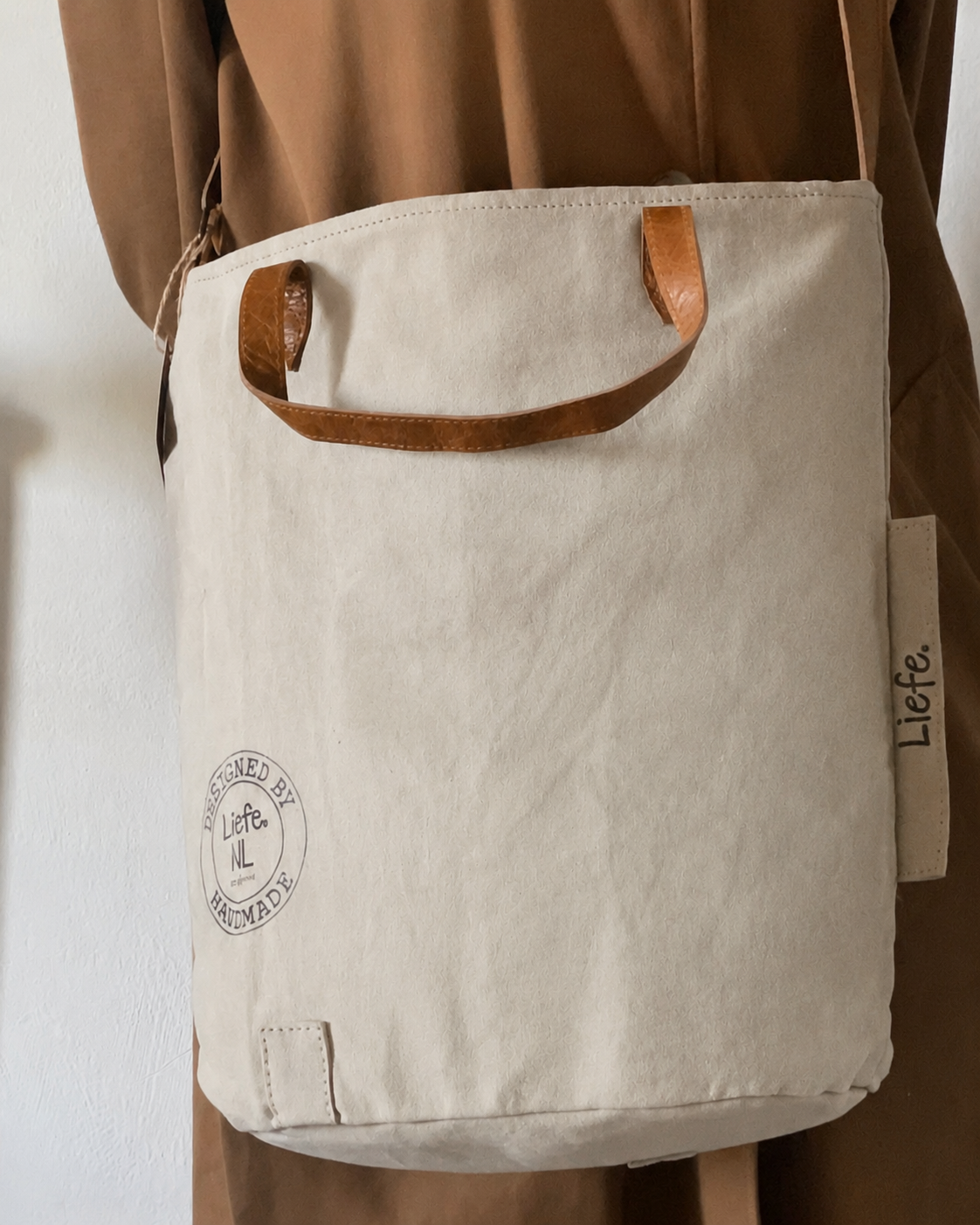 A canvas tote bag in a warm off-white color with brown leather handles, worn crossbody by a person wearing a brown coat. The bag features a circular 'Lief & Lief' logo on the lower left and a side tag with the brand name 'Lief & Lief'.