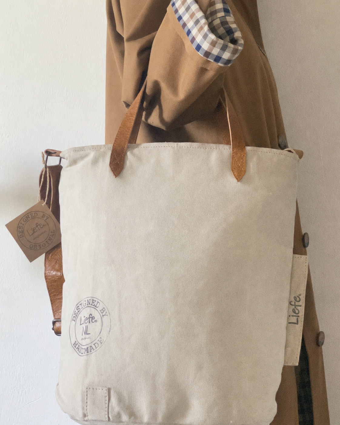 A canvas tote bag with brown leather handles and a matching leather strap tag, hanging against a light wall next to a brown coat. The bag has a circular logo printed on the front and the brand name 'Lifa' visible on a side tag.