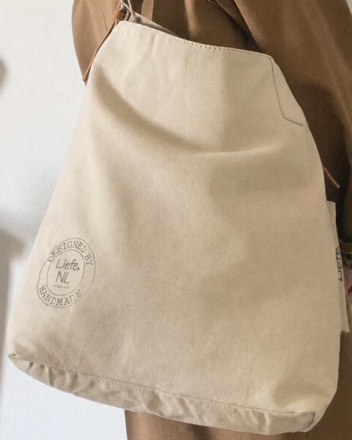 A beige canvas tote bag with a circular 'Liefte NL' logo printed on the front, shown being worn over the shoulder of someone wearing a brown jacket.