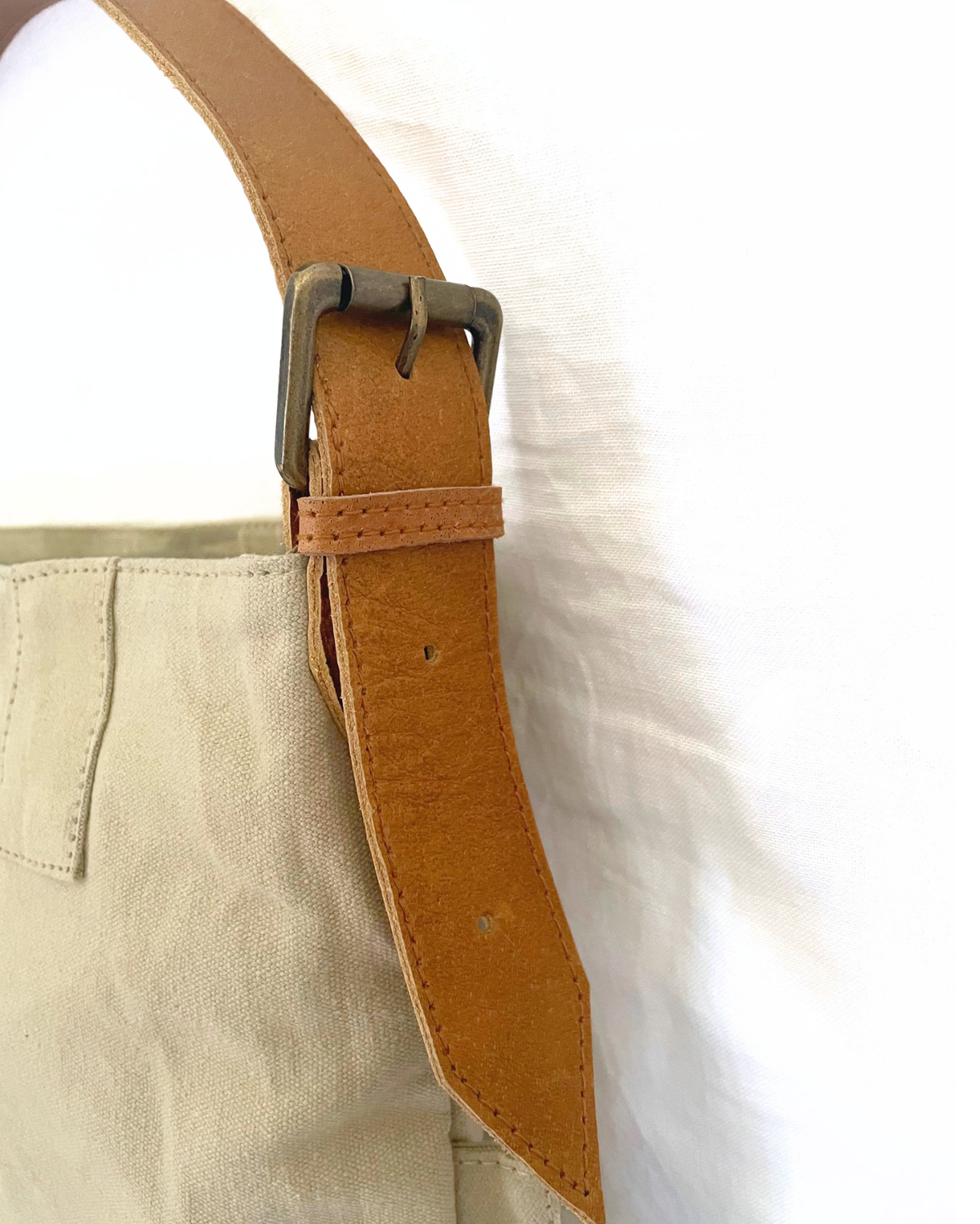 Close-up of a tan leather belt with a metal buckle attached to a light beige canvas bag, set against a white fabric background.