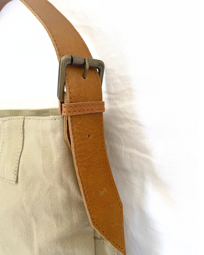 Close-up of a tan leather belt with a metal buckle attached to a light beige canvas bag, set against a white fabric background.