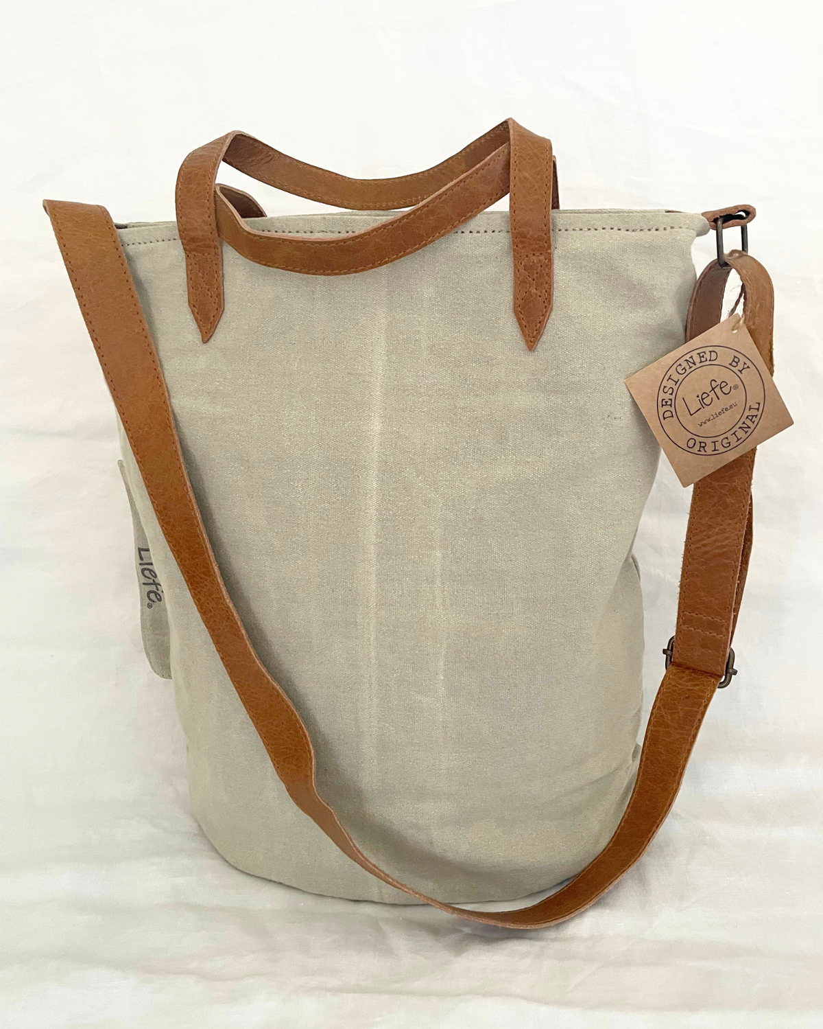 A light beige canvas tote bag with brown leather handles, a matching adjustable shoulder strap, and a hanging leather brand tag that reads 'DESIGNED BY LIFE TOUGH'.