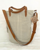 A light beige canvas tote bag with brown leather handles, a matching adjustable shoulder strap, and a hanging leather brand tag that reads 'DESIGNED BY LIFE TOUGH'.