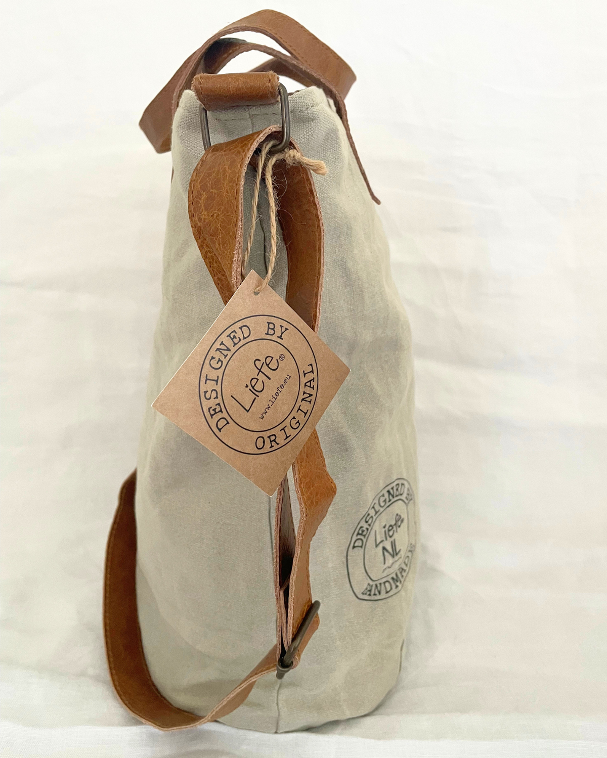 A canvas bucket bag in a light gray color with brown leather straps and a hanging paper tag that reads 'DESIGNED BY J&L ORIGINAL'.