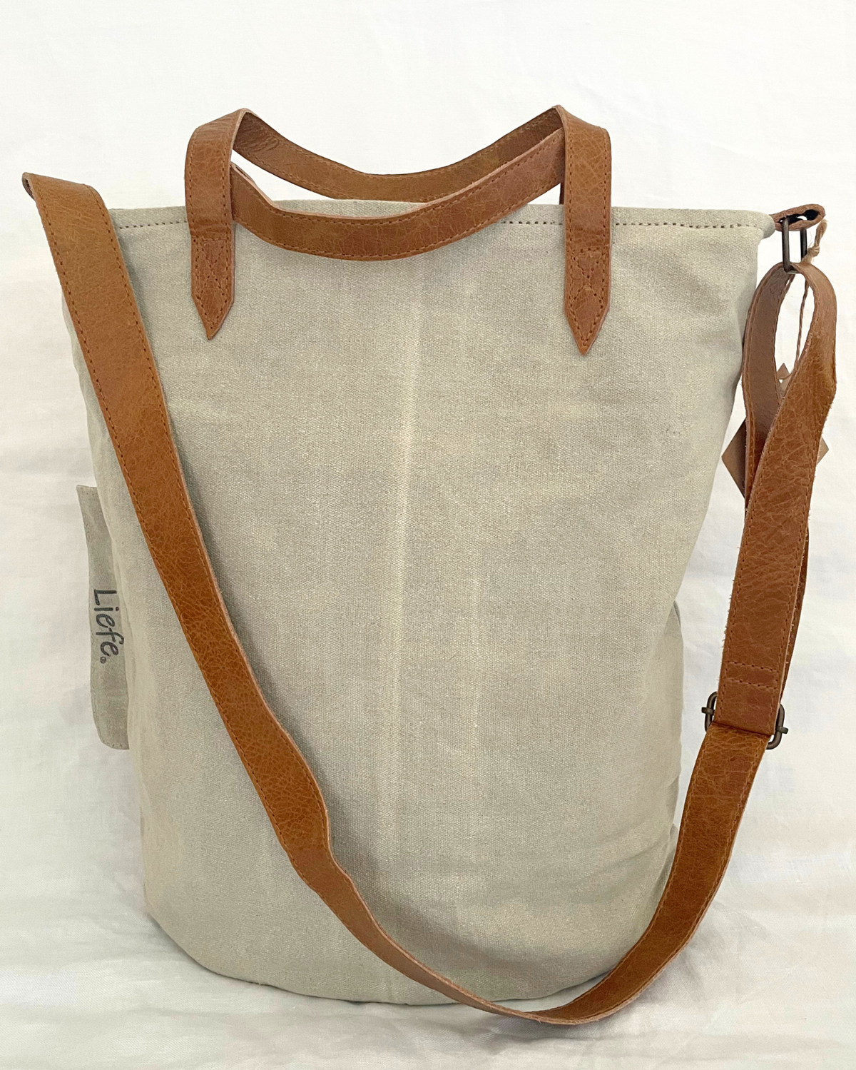 A light beige canvas bucket tote bag with brown leather handles and a matching adjustable shoulder strap, shown against a white fabric background.